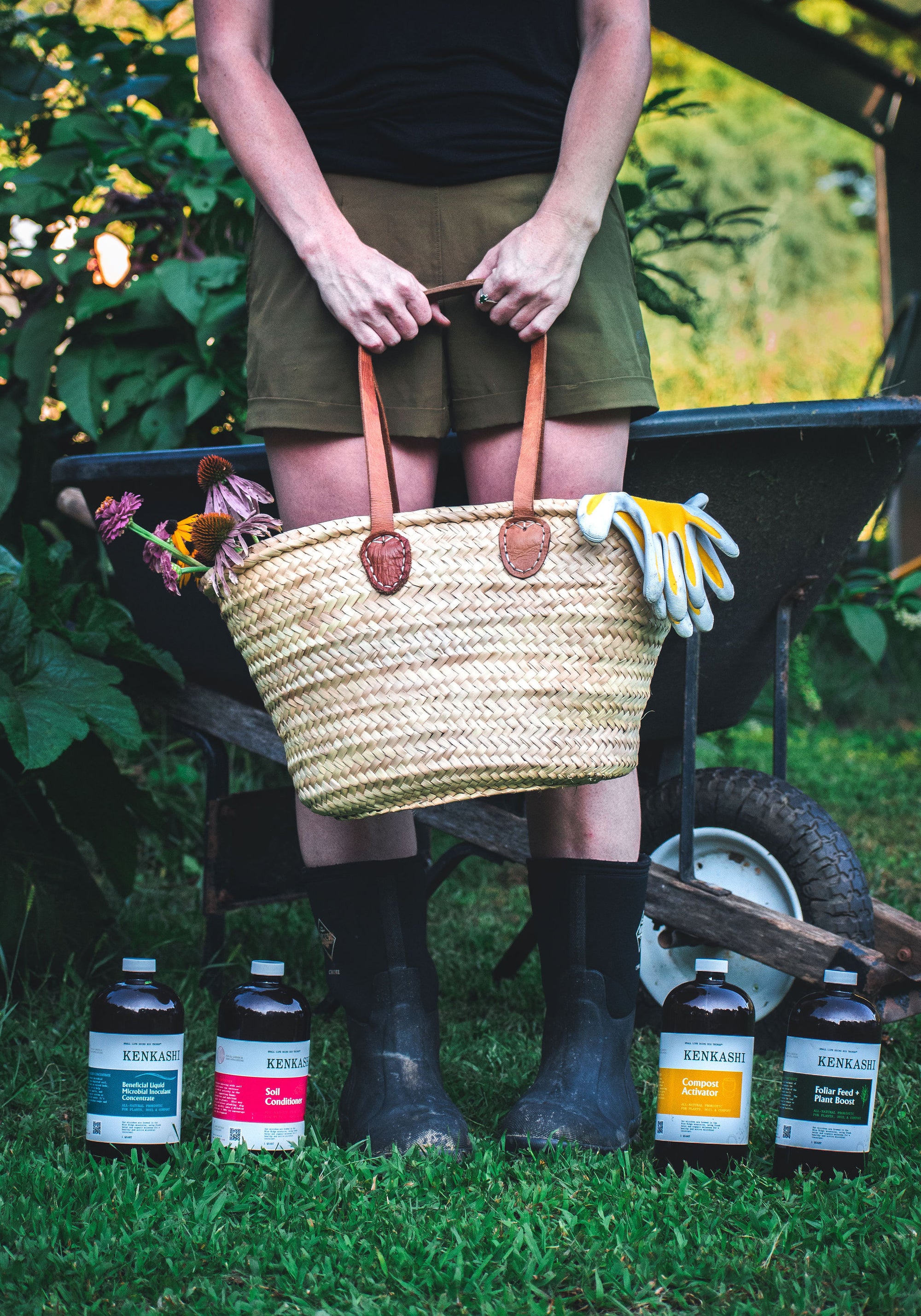 Woman holding a basket with gloves and cut flowers next to Kenkashi liquid microbial inoculant bottles in the grass