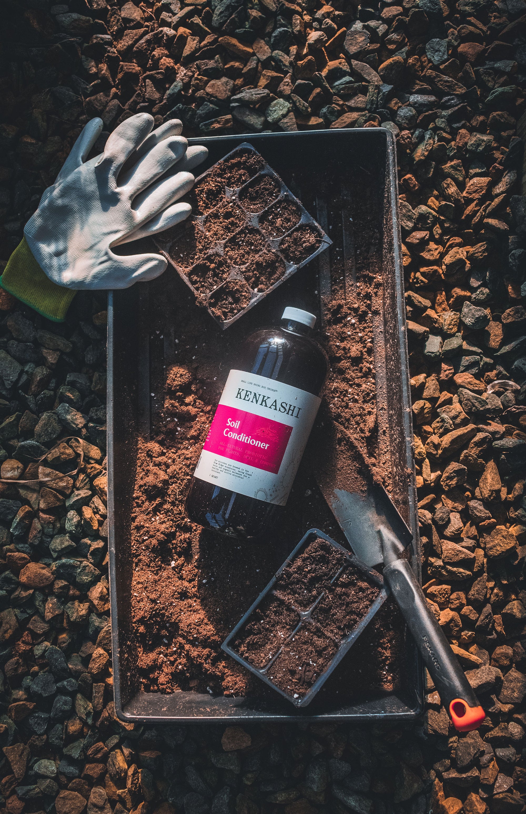 Bottle of Kenkashi soil conditioner liquid inoculated microbial concentrate on a tray of seed starter soil shovel and gloves 
